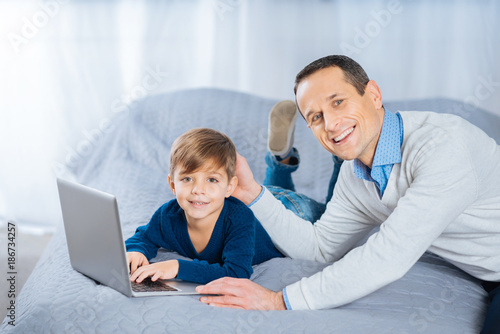 Pure affection. Upbeat pleasant man lying on the bed next to his son and patting his head fondly while the boy chatting with his friends online, using laptop