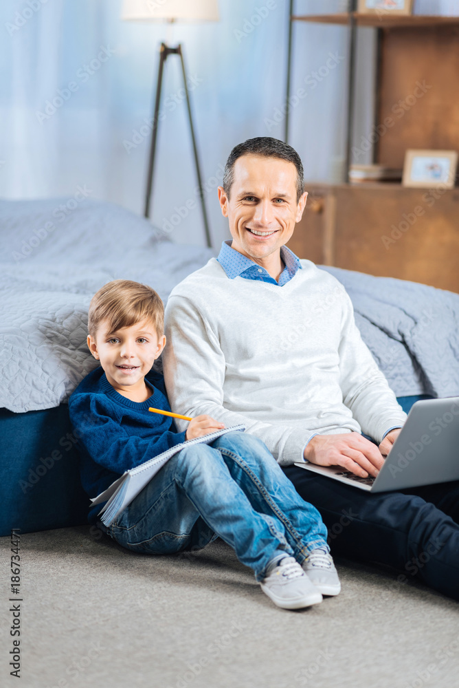 Different hobbies. Pleasant young man sitting on the bedroom floor and using laptop while his little son sitting next to him and drawing in the drawing book Different hobbies. Pleasant young man sitting on the bedroom floor and using laptop while his little son sitting next to him and drawing in the drawing book