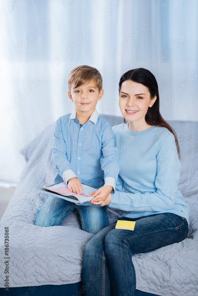 Mother-son bond. Pleasant young woman and her little son sitting on the bed together and posing for the camera while sticking memos to the notebook Mother-son bond. Pleasant young woman and her little son sitting on the bed together and posing for the camera while sticking memos to the notebook