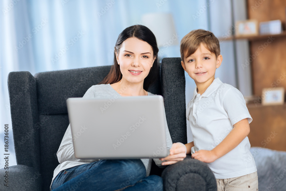 Nice pastime. Upbeat young woman and her little son watching a video on the laptop together and posing for the camera Nice pastime. Upbeat young woman and her little son watching a video on the laptop together and posing for the camera