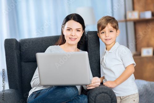 Nice pastime. Upbeat young woman and her little son watching a video on the laptop together and posing for the camera