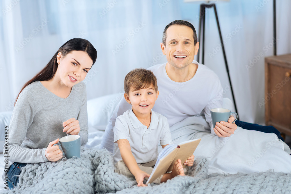 Sweet care. Sweet young family sitting on the bed and posing for the camera while the boy reading the book to his parents drinking coffee