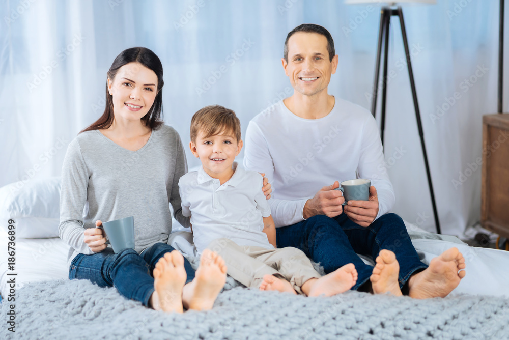 Happy moments. Beautiful young family sitting on the bed, holding tea cups and posing for the camera, smiling brightly Happy moments. Beautiful young family sitting on the bed, holding tea cups and posing for the camera, smiling brightly