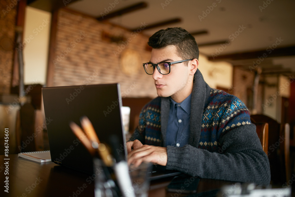 Handsome freelancer businessman in glasses and winter sweater diligently working on laptop in cafe. Man typing on keyboard and searches new job on internet at coffeeshop. Business concept Handsome freelancer businessman in glasses and winter sweater diligently working on laptop in cafe. Man typing on keyboard and searches new job on internet at coffeeshop. Business concept
