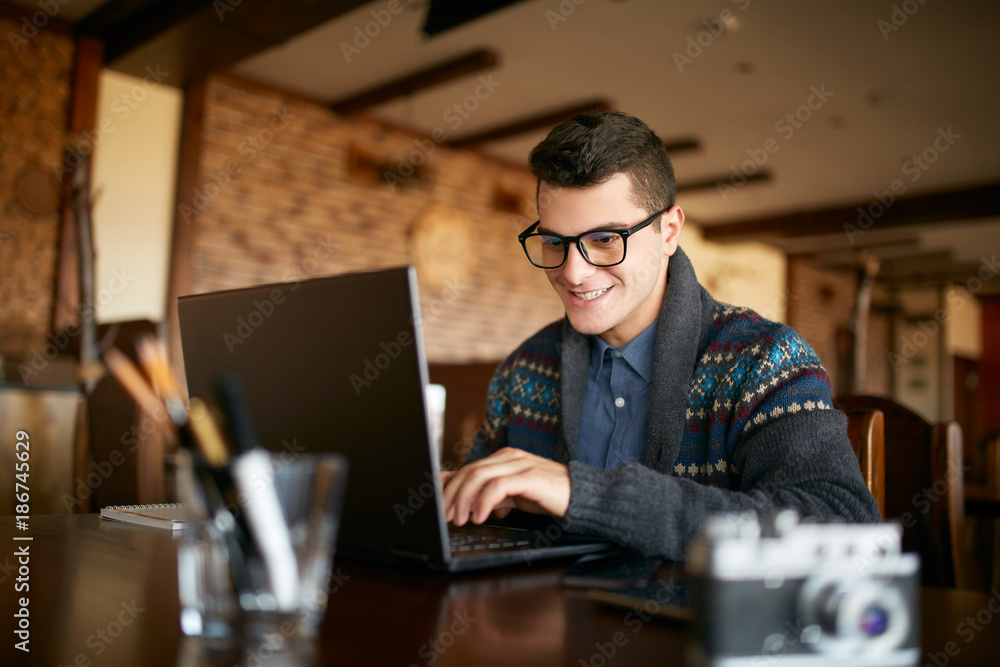 Smiling attractive hipster man in glasses and winter sweater working on laptop in cafe. Freelancer businessman browsing internet at coffeshop. Photographer retouching photos. Vintage camera on table. Smiling attractive hipster man in glasses and winter sweater working on laptop in cafe. Freelancer businessman browsing internet at coffeshop. Photographer retouching photos. Vintage camera on table.