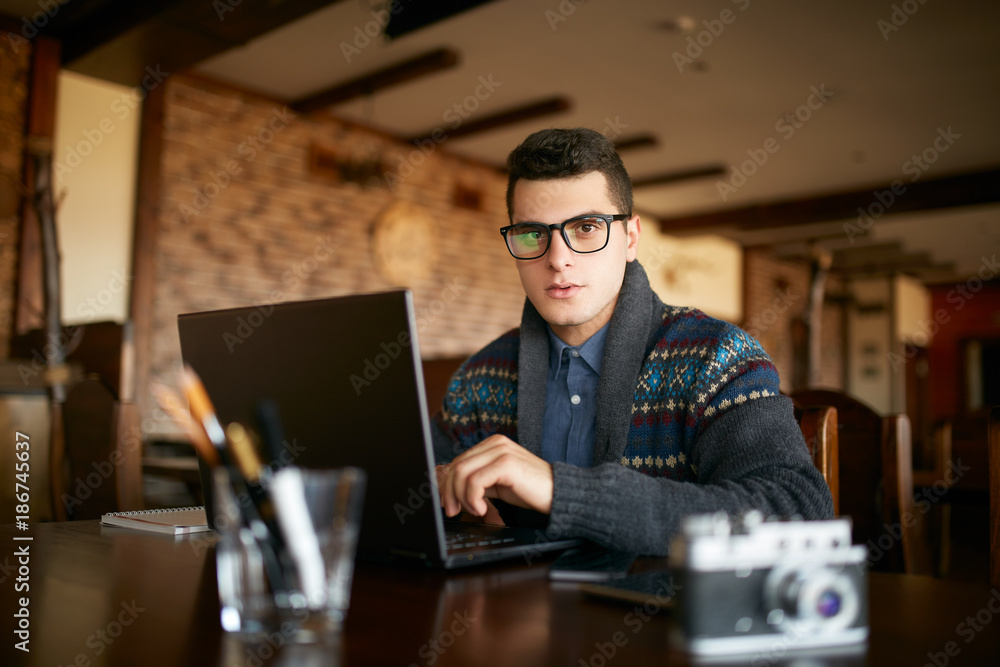 Authentic portrait of young confident businessman looking at camera with laptop in office. Hipster man in glasses and knitted sweater doing his startup ICO project. Smart casual wear. Business theme Authentic portrait of young confident businessman looking at camera with laptop in office. Hipster man in glasses and knitted sweater doing his startup ICO project. Smart casual wear. Business theme