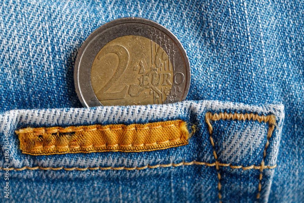 Euro coin with a denomination of two euro in the pocket of worn old blue denim jeans with orange seam Euro coin with a denomination of two euro in the pocket of worn old blue denim jeans with orange seam