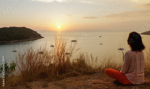 Young girl sitting in yoga lotus meditation position in front to seaside on the rocks an watching the sun goes down in a golden hour. sunset over sea.