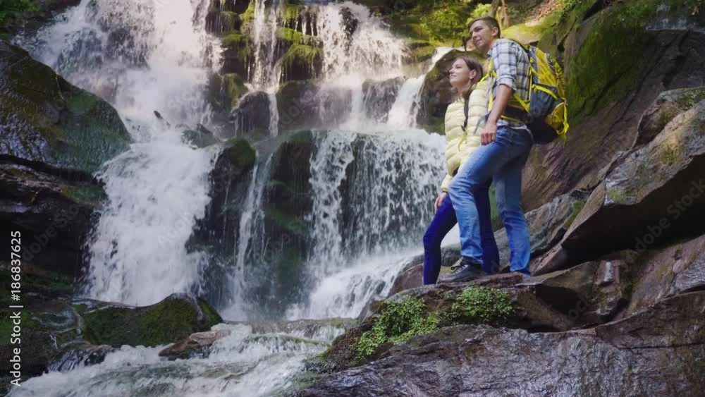 A young couple of tourists with backpacks admiring the waterfall in the mountains. Stand on slippery stones A young couple of tourists with backpacks admiring the waterfall in the mountains. Stand on slippery stones