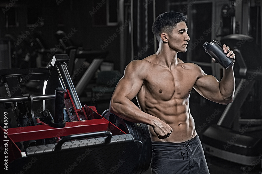 Naklejka premium A young handsome, well-trained Asian man, in the gym, after trimming the biceps, triceps, legs, back, resting and drinking protein from the shaker.