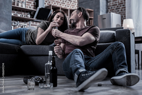 Alcohol addicted couple. Happy cheerful bearded man smiling and giving his wife a drink while sitting on the floor