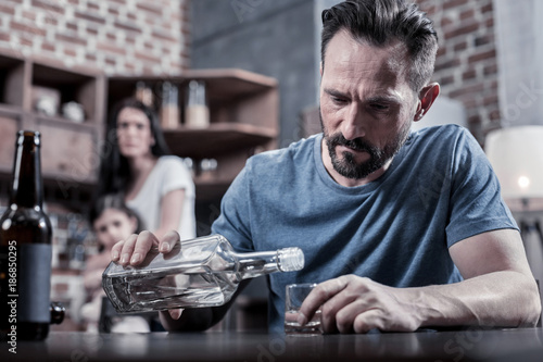 Drinking father. Serious unhappy sad man sitting at the table and pouring vodka into his glass while having alcohol addiction photo