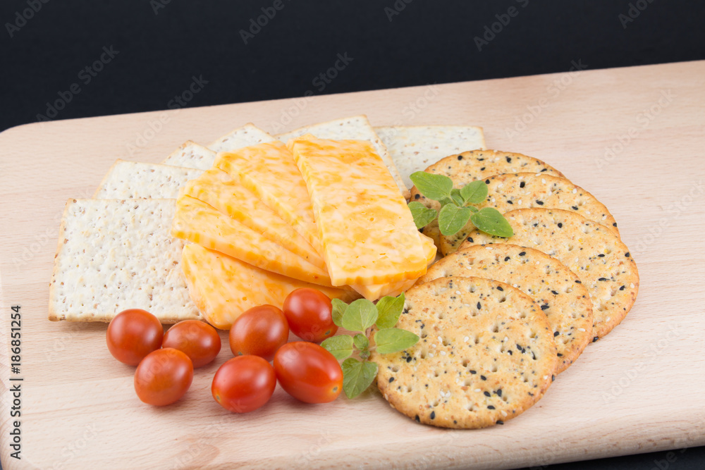Sliced cheese with crackers fresh tomatoes and oregano on a wooden cutting board with a black background Sliced cheese with crackers fresh tomatoes and oregano on a wooden cutting board with a black background