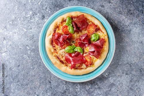 Traditional pizza with bresaola, cheese, tomatoes and basil served on turquoise plate over blue texture background. Top view with space