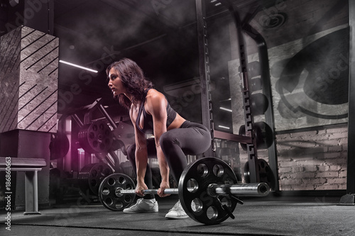 Young beautiful muscular girl sportsman bodybuilder doing exercises in a modern gym using a barbell, against a dark background.