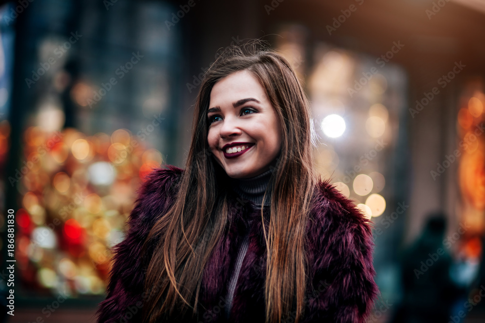 Portrait pretty girl with long hair in Burgundy fur coat of artificial fur knitted hat and bright burgundy lips walking on street with coffee to go. City lifestyle