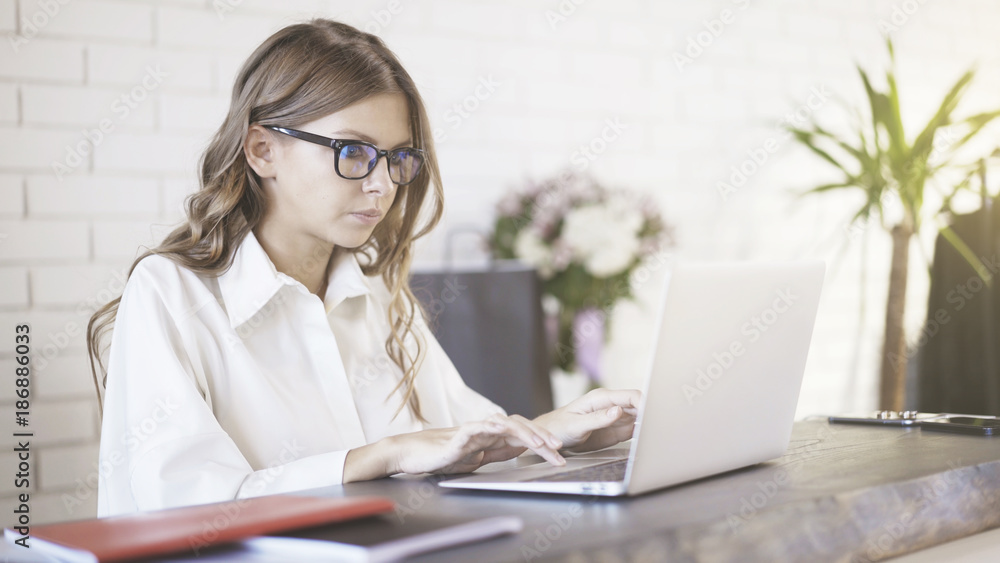Side view of a young beautiful woman wearing glasses typing at her laptop in office. Portrait shot. Side view of a young beautiful woman wearing glasses typing at her laptop in office. Portrait shot.