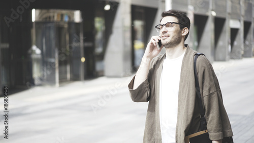 A hipster guy wearing glasses and a khaki jacket talking on his smartphone and laughing in the street. Portrait shot