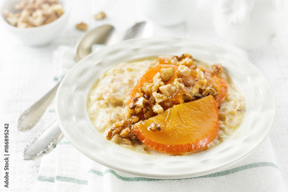 Oatmeal porridge with caramelized persimmon and walnuts in a light ceramic bowl on a white wooden background. Selective focus. Top view. Copy space. Oatmeal porridge with caramelized persimmon and walnuts in a light ceramic bowl on a white wooden background. Selective focus. Top view. Copy space.