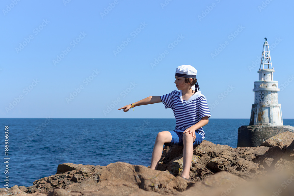 A boy in a striped T-shirt and cap sits on the rocks and looks at the lighthouse.