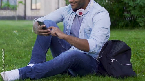 Mixed-race guy sitting on grass with mobile phone and headphones, application photo