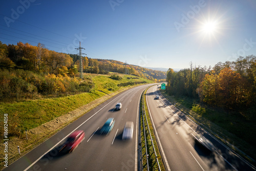 Motion blurred cars on the highway surrounded by forest in autumn colors with sun shining in the sky. View from above.