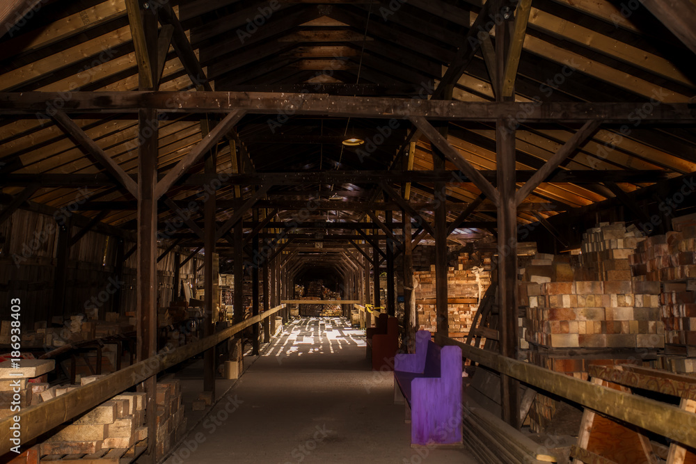 Wood shell of rafters and beams dotted with sunlight storing stacks of bricks between a hallway of a discontinued brick factory