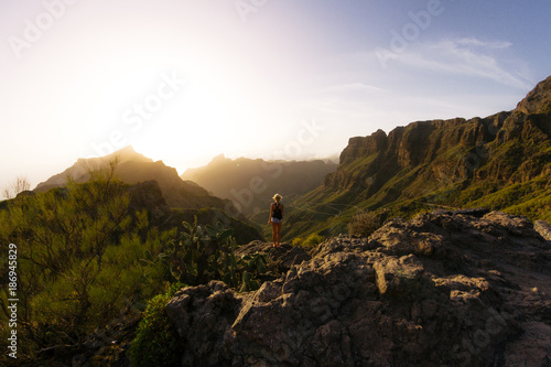 Young blond woman in shorts looking at rocky landscape with steep cliffs while sunset