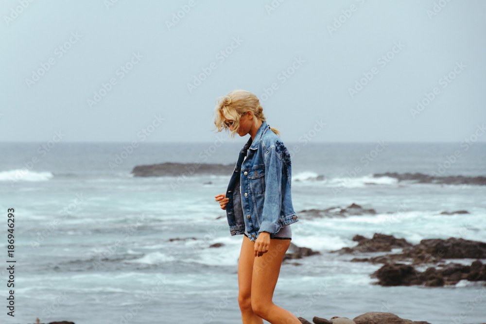 Young fashion woman in jeans jacket and mini shorts posing on coastline with ocean waves in background