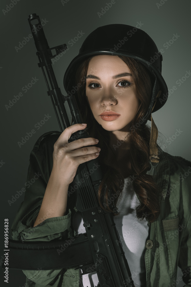 beautiful young woman in military helmet with rifle, on grey with shadows