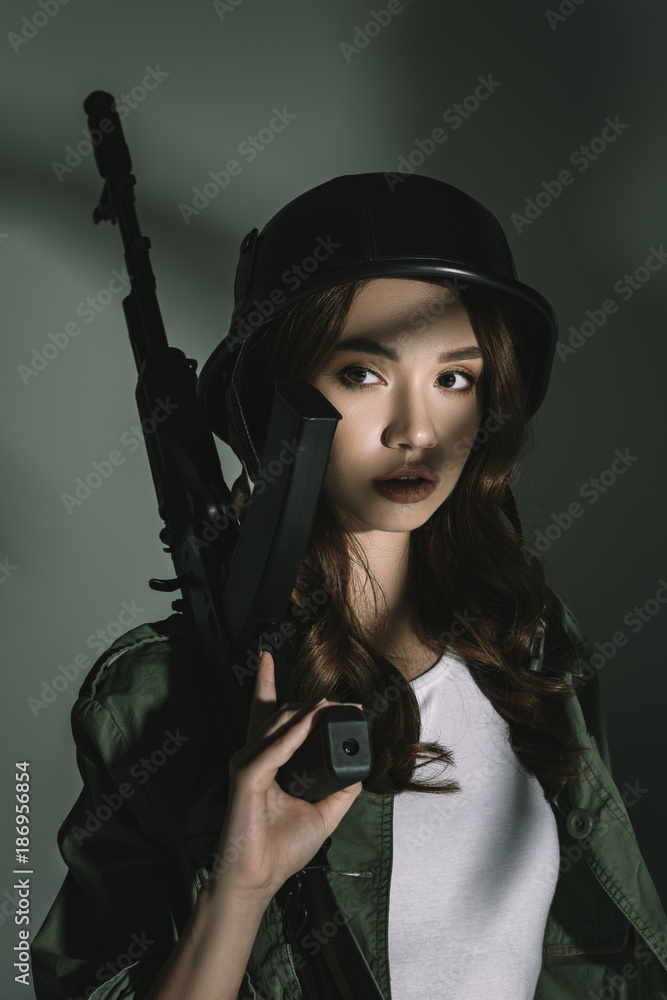 beautiful girl posing in military helmet with rifle, on grey with shadows beautiful girl posing in military helmet with rifle, on grey with shadows