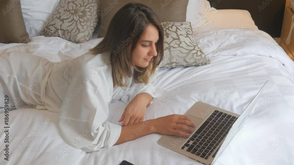 Young woman in bathrobe lying on bed and working on laptop Young woman in bathrobe lying on bed and working on laptop