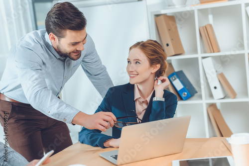 standing man in blue shirt speaking to sitting woman against laptop on table