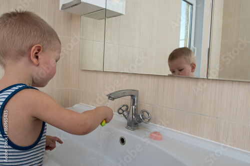 Happy boy taking bath in kitchen sink. Child playing with foam and soap bubbles in sunny bathroom with window. Little baby bathing. Water fun for kids. Hygiene and skin care for children. 