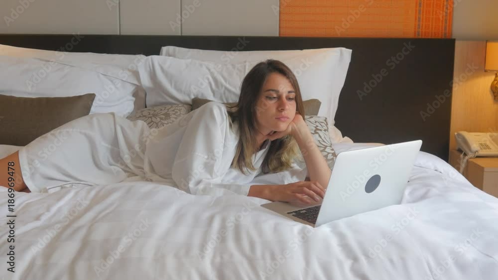 Young woman in bathrobe lying on bed and working on laptop Young woman in bathrobe lying on bed and working on laptop