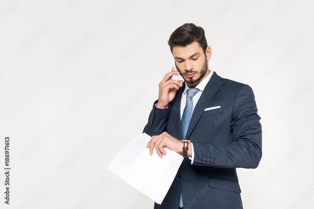 young businessman holding papers and talking on smartphone while checking wristwatch isolated on grey young businessman holding papers and talking on smartphone while checking wristwatch isolated on grey