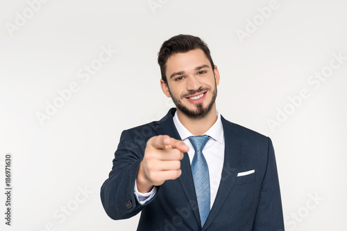 smiling young businessman pointing at camera isolated on grey