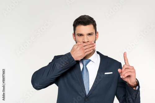 young businessman closing mouth with palm and pointing up with finger isolated on grey