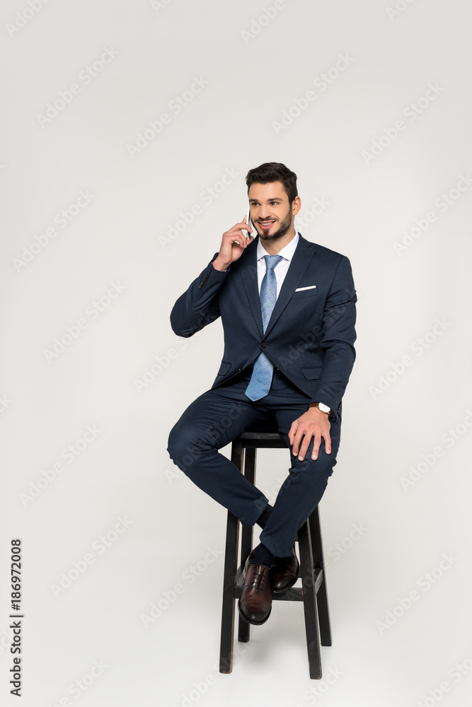smiling young businessman talking on smartphone while sitting on stool and looking away isolated on grey smiling young businessman talking on smartphone while sitting on stool and looking away isolated on grey