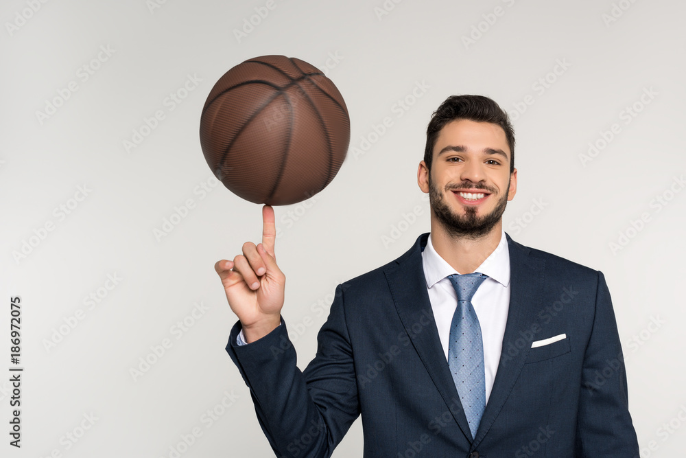 young businessman holding basketball ball on finger and smiling at camera isolated on grey