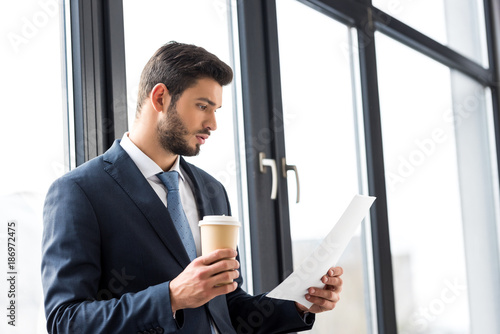 young businessman holding coffee to go and reading papers in office