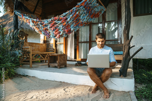 Work home. Handsome young man using laptop sitting on the doorstep of his house on a hammock background.