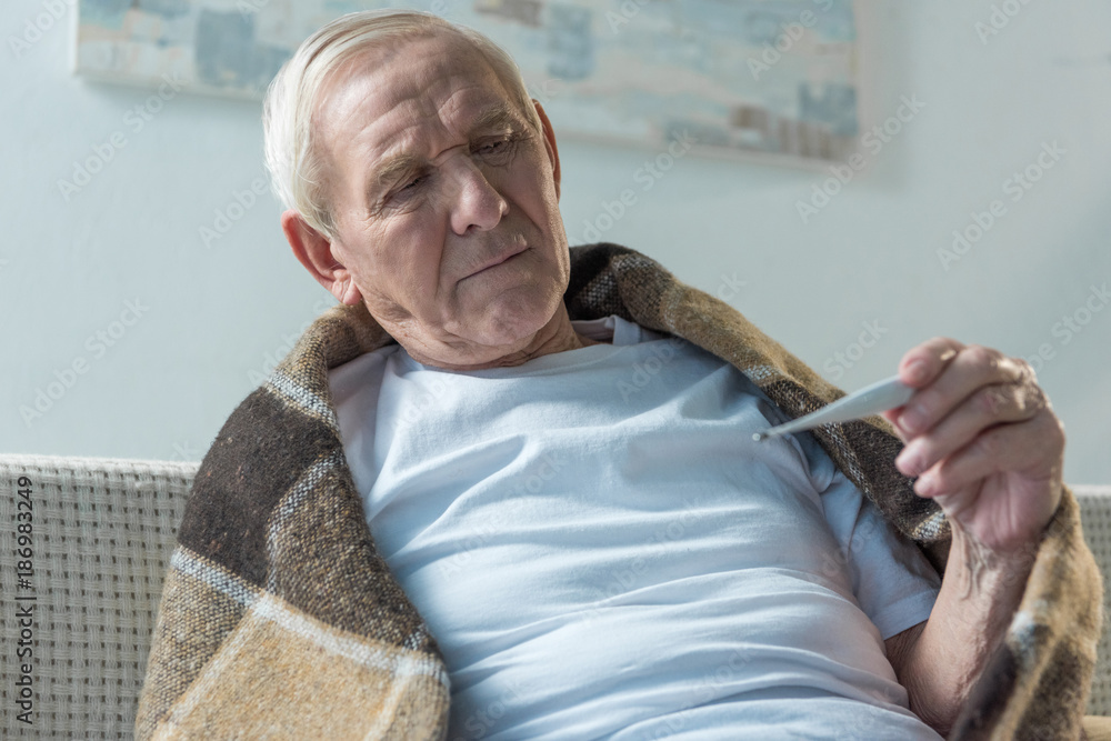 Senior sick man covered in plaid sitting on sofa and checking thermometer Senior sick man covered in plaid sitting on sofa and checking thermometer