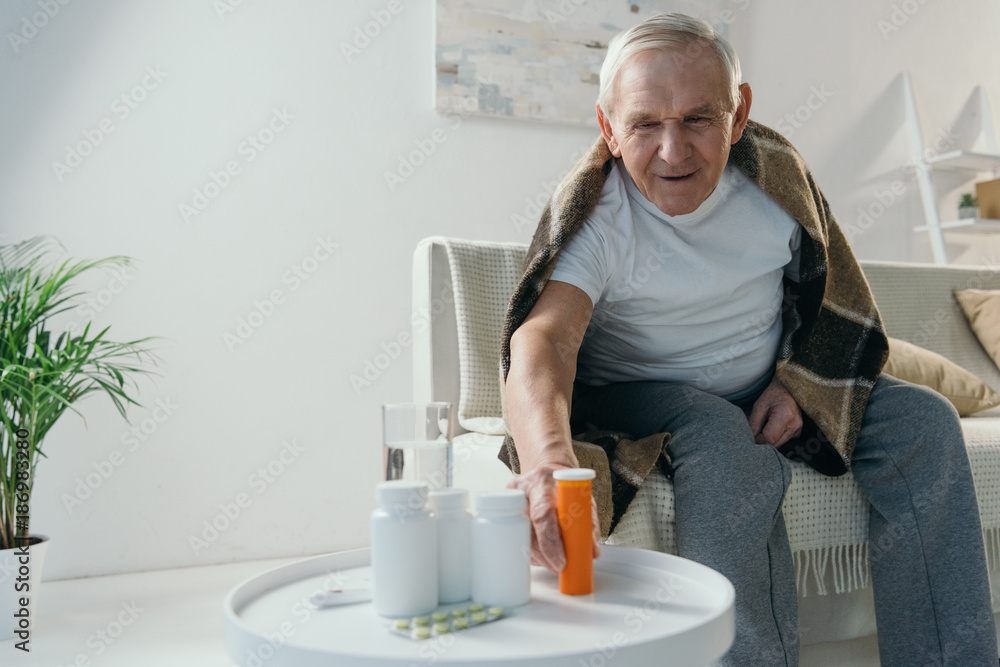 Senior sick man covered in plaid takes medications from table Senior sick man covered in plaid takes medications from table