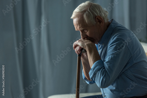 Fototapeta Senior sad man leans on a cane while sitting on sofa