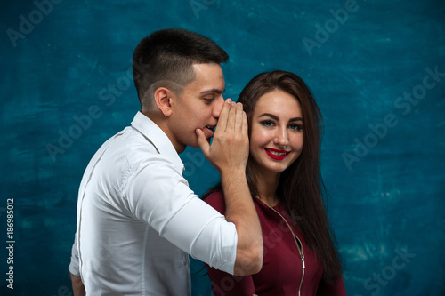 Loving couple - young man whispering something in the ear of his beautiful woman on blue background