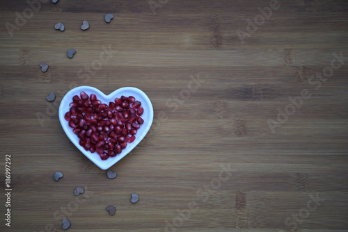 healthy food photography image of ripe juicy shiny red pomegranate seeds in a white love heart shape dish on a natural dark wood background with copy space