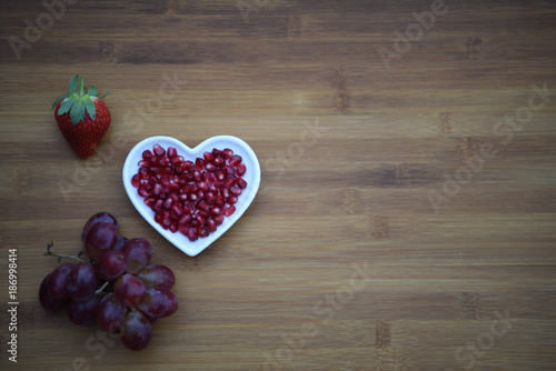 food photography image with red shiny pomegranate seeds in a white love heart shape dish with a strawberry and bunch of fresh purple grapes on a dark natural wood background with copy space