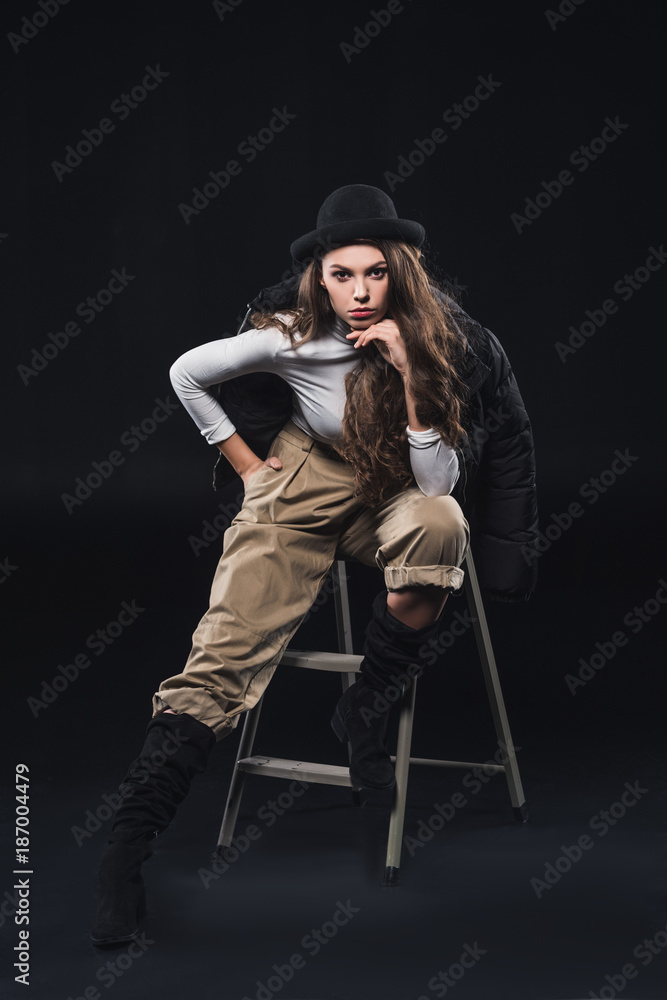 beautiful young woman in fashionable hat and winter jacket sitting on ladder and looking at camera on black