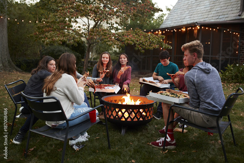 Teenage friends sit round a fire pit eating take-away pizza